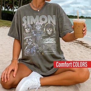 May include: A gray t-shirt featuring a graphic of a skull-faced character with the name "SIMON Riley" above. The shirt also has the text "CENTRAL POLICE DEPT" and "SIMON GHOST RILEY". The shirt is labeled "Comfort COLORS".