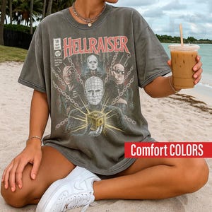 May include: A charcoal gray short-sleeve t-shirt featuring a graphic print of the movie Hellraiser. The design includes the text HELLRAISER, 18 SEP 1987, and $1.50, along with illustrations of characters and a puzzle box surrounded by chains.