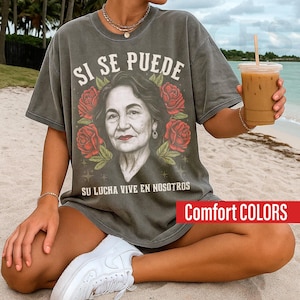 May include: A gray t-shirt with the text "SI SE PUEDE" above a portrait of a woman surrounded by red roses. The phrase "SU LUCHA VIVE EN NOSOTROS" is below the portrait. The shirt has the words "Comfort COLORS" in a red rectangle.