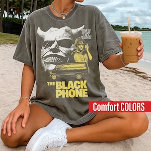 May include: Dark gray t-shirt featuring a graphic from "The Black Phone" movie. The design includes a monster face, a boy on a yellow car, and the movie title in yellow. The shirt is labeled "Comfort COLORS".