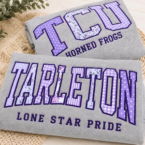 May include: Two folded grey sweatshirts featuring purple applique lettering. The top sweatshirt displays TCU HORNED FROGS, while the bottom sweatshirt displays TARLETON LONE STAR PRIDE. The letters feature various purple and white patterns.