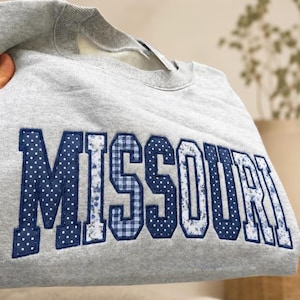May include: A light gray crewneck sweatshirt featuring the word MISSOURI appliqued across the chest in large block letters. The letters are crafted from various blue and white fabrics, including polka dot, floral, and gingham patterns.