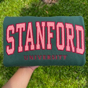 May include: A dark green sweatshirt featuring the word STANFORD in large, pink, polka-dotted block letters with a dark red outline. Below the word STANFORD, the word UNIVERSITY is embroidered in smaller, dark red, capital block letters.