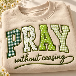 May include: A beige crewneck sweatshirt featuring the word PRAY in large, block-style letters with varied green patterns including gingham, floral, and polka dots. Below the word PRAY, the phrase without ceasing is embroidered in dark green script.