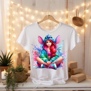 May include: White t-shirt featuring a colorful illustration of a character with red hair, wearing a blue hooded jacket and pink sneakers. The design includes watercolor-style splashes of pink, blue, and green.