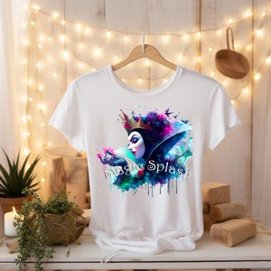 May include: White t-shirt featuring a watercolor design of a queen with a crown, surrounded by colorful splashes and the text "Magic Splash". The design includes shades of purple, blue, and green.