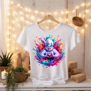 May include: White t-shirt featuring a colourful graphic of Ursula from The Little Mermaid making a heart shape with her hands. The graphic is surrounded by colourful paint splatters.