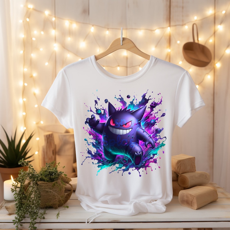 May include: White t-shirt with a graphic print of a purple and blue cartoon ghost character with red eyes. The ghost is surrounded by colorful paint splatters.