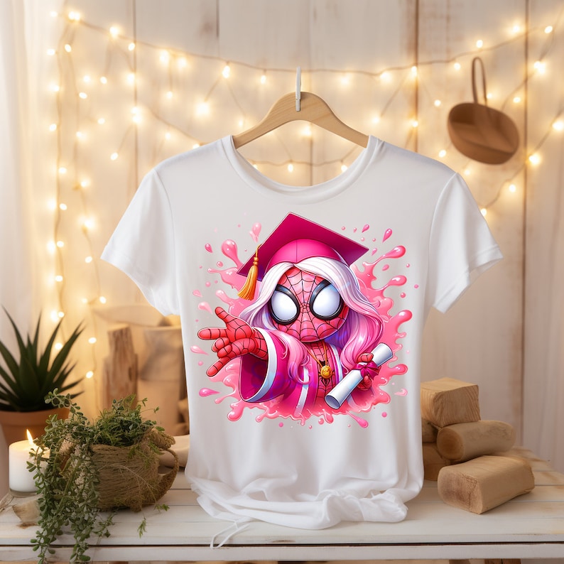 May include: White t-shirt with a pink and red cartoon image of a Spider-Man character wearing a graduation cap and gown. The character is holding a diploma and is surrounded by pink paint splatters.