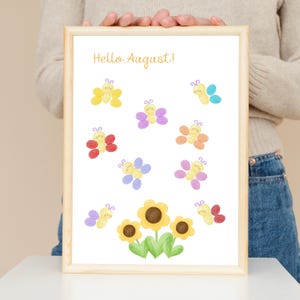 Puede incluir: Una l&aacute;mina enmarcada con el texto Hello August en letra cursiva naranja en la parte superior. Debajo del texto hay ocho peque&ntilde;as mariposas coloridas con caras sonrientes, dispuestas alrededor de tres girasoles amarillos con centros marrones y hojas verdes.