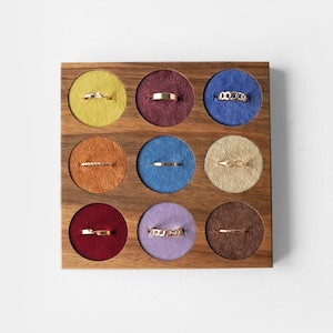 May include: A square wooden ring display organizer featuring nine circular cutouts lined with colorful velvet fabric. Each cutout holds a single gold-toned metal ring. The velvet colors include yellow, maroon, blue, orange, light blue, beige, red, lavender, and brown.