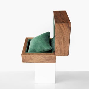May include: A wooden watch box with a hinged lid, open to reveal a green velvet pillow. The interior is lined with green fabric. The box is set on a white base, creating a clean, modern aesthetic.
