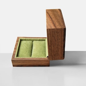May include: A wooden jewelry box with a hinged lid, open to reveal a light green velvet interior with two compartments. The box is made of dark brown wood and sits on a white surface.