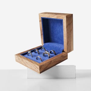 May include: A wooden ring box with a blue velvet interior, open to display several rings. The box is made of light brown wood and is resting on a white surface. The rings are silver and gold.