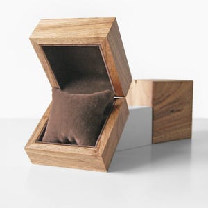 May include: Wooden watch box with a hinged lid, open to reveal a brown velvet pillow. The box is made of light-colored wood with visible grain. A separate wooden cube is positioned next to the open box, suggesting additional storage or design.