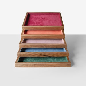 May include: A stack of five wooden trays with velvet interiors in various colors: red, peach, pink, blue, and green. The trays are rectangular with a raised wooden border, showcasing a minimalist design.
