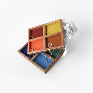 Puede incluir: Un organizador de joyas de madera de dos niveles con múltiples compartimentos cuadrados forrados de terciopelo en varios colores, incluyendo rojo, naranja, amarillo, azul y verde. Varios anillos dorados y una cadena de oro están colocados dentro.
