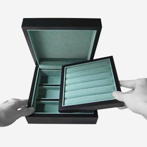 May include: A black wooden jewelry box with a hinged lid and a removable tray. The interior is lined with a light green fabric. The box is open, and a hand is lifting the tray.
