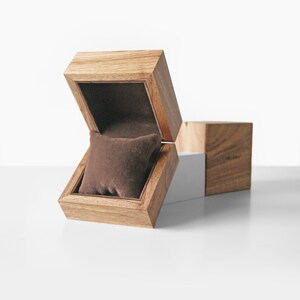 May include: Wooden watch box with a hinged lid, open to reveal a brown velvet pillow. The box is made of light-colored wood with visible grain. A separate wooden cube is positioned next to the open box, suggesting additional storage or design.