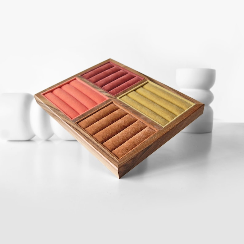 May include: A square wooden jewelry display tray featuring four distinct sections lined with velvet. Each section contains four padded slots in varying colors: coral, deep red, mustard yellow, and brown, designed for organizing rings.
