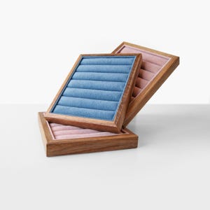 May include: Three wooden jewelry trays with velvet inserts. One tray has a blue insert, and two have pink inserts. The trays are rectangular with a grooved design in the velvet. The wooden frames have a natural wood grain.