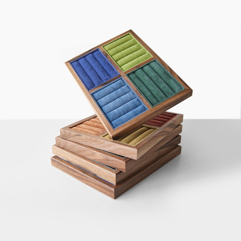 May include: A stack of square wooden jewelry display trays with velvet-lined inserts. The top tray is angled, revealing four quadrants in blue, light green, dark green, and light blue. Lower trays feature inserts in orange, red, and gold tones.