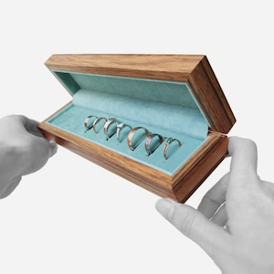 May include: A wooden ring box with a light blue velvet interior. The box is open and contains eight rings.