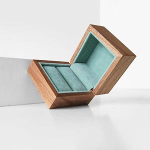 Puede incluir: Una caja de madera para anillos con un interior de terciopelo azul claro. La caja está abierta y la tapa está levantada.