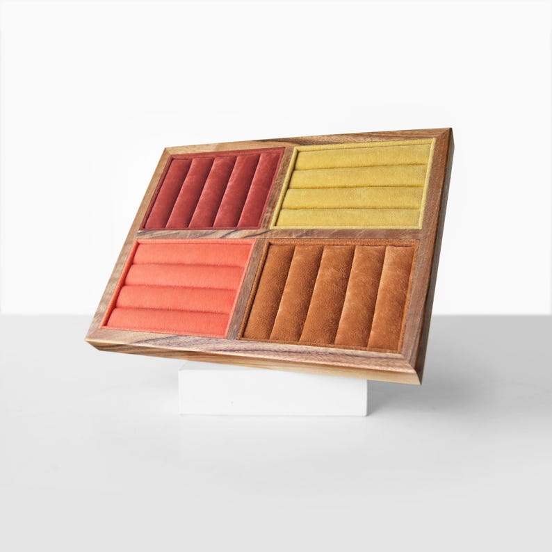 May include: A rectangular wooden jewelry display tray featuring four distinct sections with velvet-lined ring slots. The sections are colored in shades of deep red, mustard yellow, coral orange, and burnt orange, set within a natural wood frame.