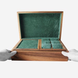Può includere: Un portagioie rettangolare in legno con finitura a venature naturali, dotato di coperchio incernierato foderato in velluto verde scuro. L'interno contiene una sezione per anelli con molteplici fessure e uno scomparto con quattro cuscinetti.