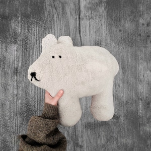 May include: A soft, plush, white stuffed animal shaped like a bear with black eyes and a black nose. The bear is being held by a hand wearing a brown sweater.