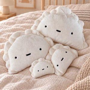 Könnte beinhalten: Ein Set aus vier weichen, weißen Plüschkissen in Form von Teigtaschen, die jeweils ein einfaches gesticktes Gesicht mit zwei schwarzen runden Augen und einem horizontalen schwarzen Strich als Mund haben, angeordnet auf beigem Stoff.