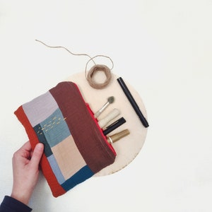 Könnte beinhalten: Eine Reißverschlusstasche aus Patchwork-Stoff mit einem roten Reißverschluss, gefüllt mit Make-up-Pinseln und Stiften. Die Tasche liegt auf einer Holzoberfläche mit einer Schnurkugel.