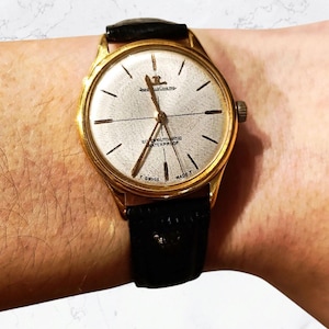 Puede incluir: Un reloj de pulsera dorado con una correa de cuero negro. La esfera del reloj es blanca con marcas negras y el texto "SUPER AUTOMATIC WATERPROOF T SWISS MADE T".