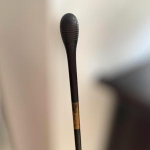 Pode incluir: Uma batuta de maestro castanho escuro com uma pega arredondada e canelada e um eixo fino. Uma faixa dourada é visível perto do meio da batuta. A batuta foi concebida para dirigir apresentações musicais.