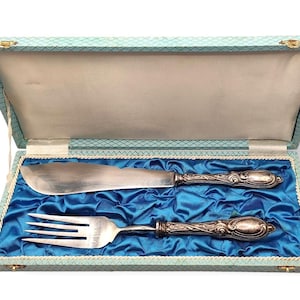 May include: A vintage butter knife and fork set in a decorative box. The silver-toned utensils have ornate handles and rest on a bed of blue satin. The box is light blue with a patterned exterior and a cream-colored interior.