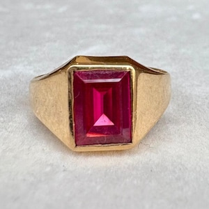 May include: A gold ring featuring a large, rectangular, vibrant red gemstone. The ring has a classic design with a simple, polished gold band and a prominent, eye-catching stone. The gemstone is approximately 0.75 inches in length.
