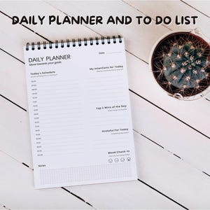 Könnte beinhalten: Ein weißes Tagesplaner-Notizbuch mit Spiralbindung, mit dem Text "DAILY PLANNER" und Abschnitten für Zeitpläne und Absichten. Ein kleiner Kaktus in einem weißen Topf befindet sich daneben auf einer weißen Holzoberfläche.