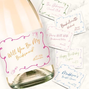 Könnte beinhalten: Eine Glasflasche mit einem weißen Etikett, das den in Gold geschriebenen Text Will You Be My Bridesmaid? in einem rosa Rahmen zeigt, begleitet von mehreren rechteckigen Karten mit verschiedenen individuellen Texten wie Bachelorette Weekend.