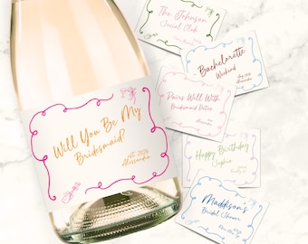 Può includere: Una bottiglia di vetro con un'etichetta bianca che riporta il testo Will You Be My Bridesmaid? in caratteri dorati all'interno di una cornice rosa, accompagnata da diverse carte rettangolari con vari testi personalizzati come Bachelorette Weekend.