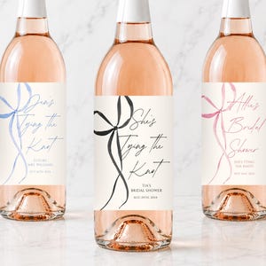 May include: Three glass wine bottles filled with pale pink rose wine, each featuring a white rectangular label. The labels display watercolor-style ribbon illustrations in blue, black, and pink, with custom text including "Tying The Knot" and dates.