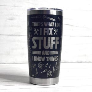 Peut inclure: Un gobelet isotherme bleu marine avec un bord et un couvercle argentés. Le gobelet est orné d'un motif argenté d'outils et du texte "THAT'S WHAT I DO I FIX STUFF AND I KNOW THINGS."