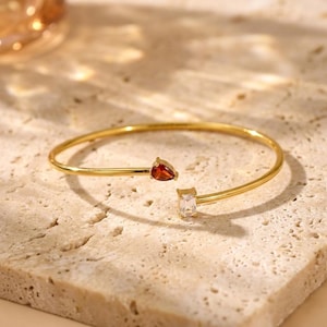 May include: A gold-colored open cuff bracelet with a teardrop-shaped red gemstone and a rectangular clear gemstone. The bracelet is displayed on a textured, light-colored surface.