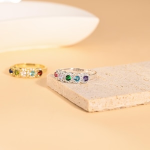 May include: Two rings with colourful gemstones. One ring is gold-toned with a row of gemstones in various colours. The other ring is silver-toned, also with a row of colourful gemstones. The rings are displayed on a neutral surface.