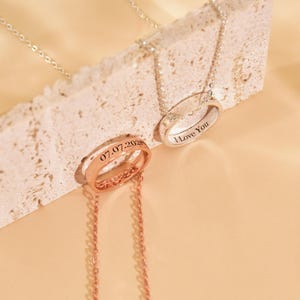 May include: Two pendant necklaces with ring-shaped charms. One is rose gold with the date "07.07.202" engraved. The other is silver with "I Love You" engraved. Both have delicate chain necklaces and are displayed on a textured surface.