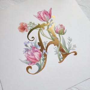 Peut inclure: Peinture à l'aquarelle de la lettre « A » dorée, ornée de tulipes roses, de petites fleurs bleues et de feuillage vert. L'œuvre est posée sur un fond blanc, créant un design floral délicat.