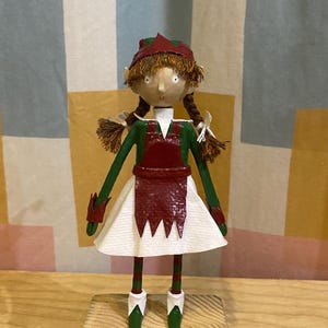 May include: A handcrafted elf doll wearing a red and green outfit. The doll features a white skirt, green arms, and green shoes. It stands on a small wooden block. The background is a patterned fabric.