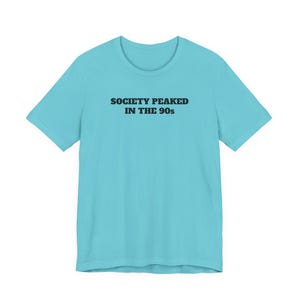 May include: A light blue t-shirt with the text "SOCIETY PEAKED IN THE 90s" printed in black. The shirt has short sleeves and a crew neck. The text is centered on the front of the shirt.