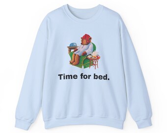 Può includere: Felpa blu chiaro con un'illustrazione di un orso di cartone animato che indossa un berretto da notte rosso e un pigiama seduto su una poltrona verde. Il testo "Time for bed." è stampato sotto l'immagine.
