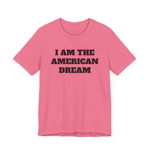 May include: Pink t-shirt with black text that reads "I AM THE AMERICAN DREAM".